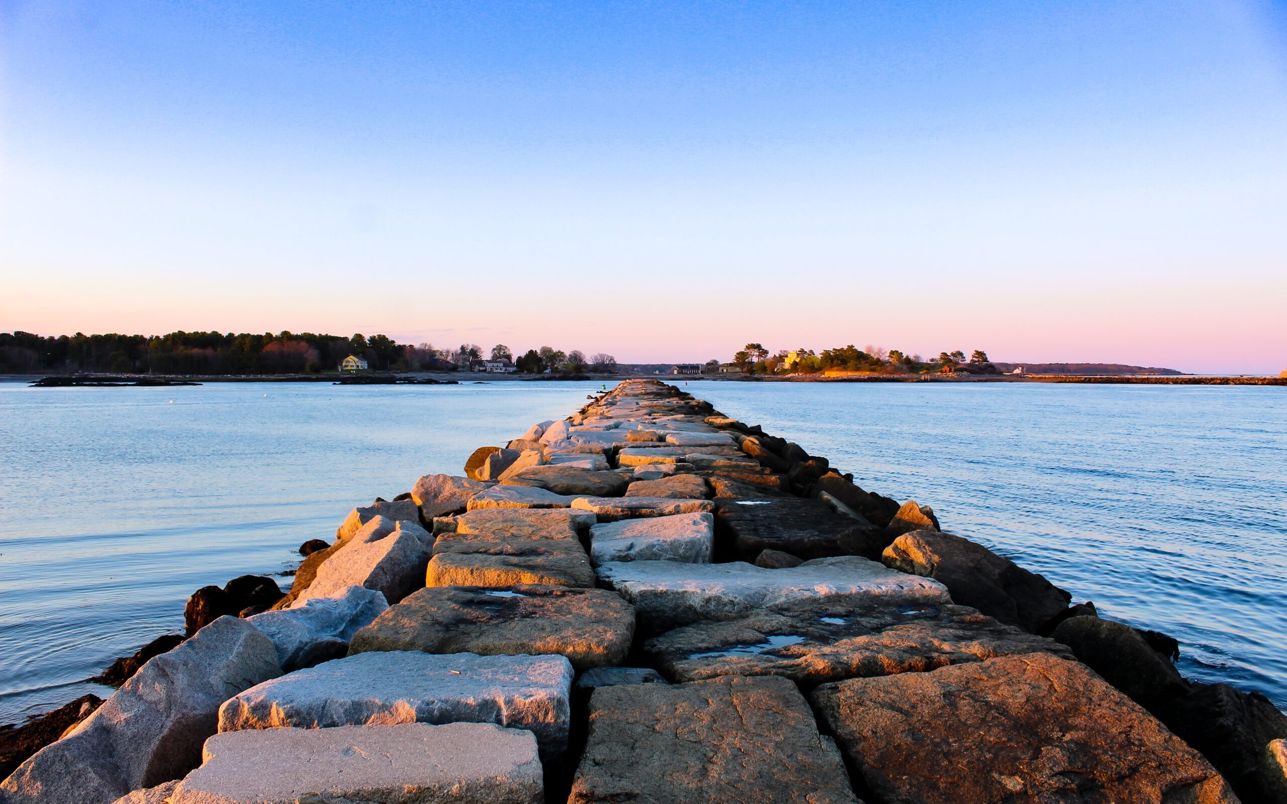 A narrow path of rocks and sea stones lead out across a bay. Across the water is land, trees, and six houses in the distance.