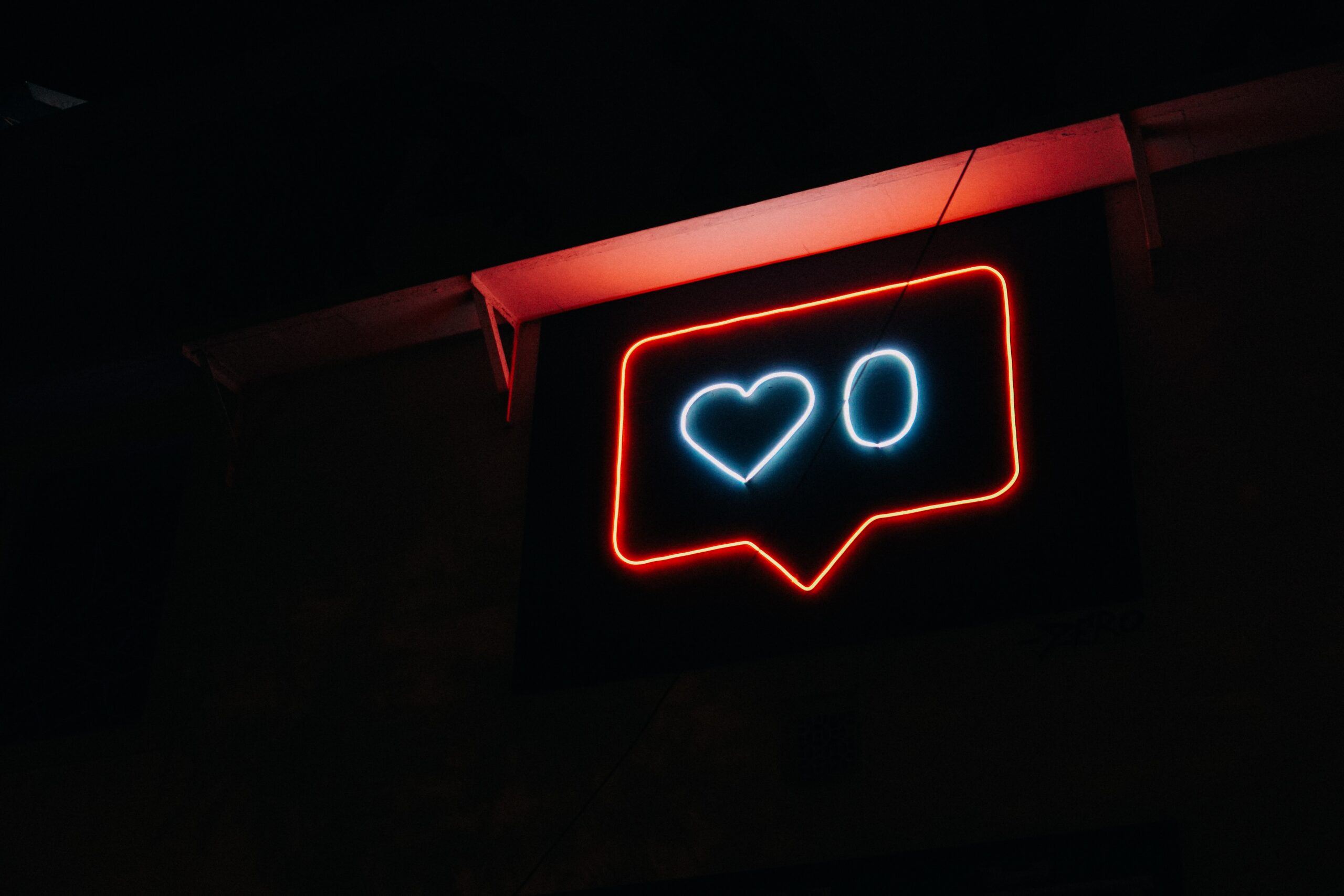 Red neon sign on in the shape of a speech bubble on a black background. A white heart next to a white zero is inside the speech bubble. The red overhang of a roof is over the speech bubble.